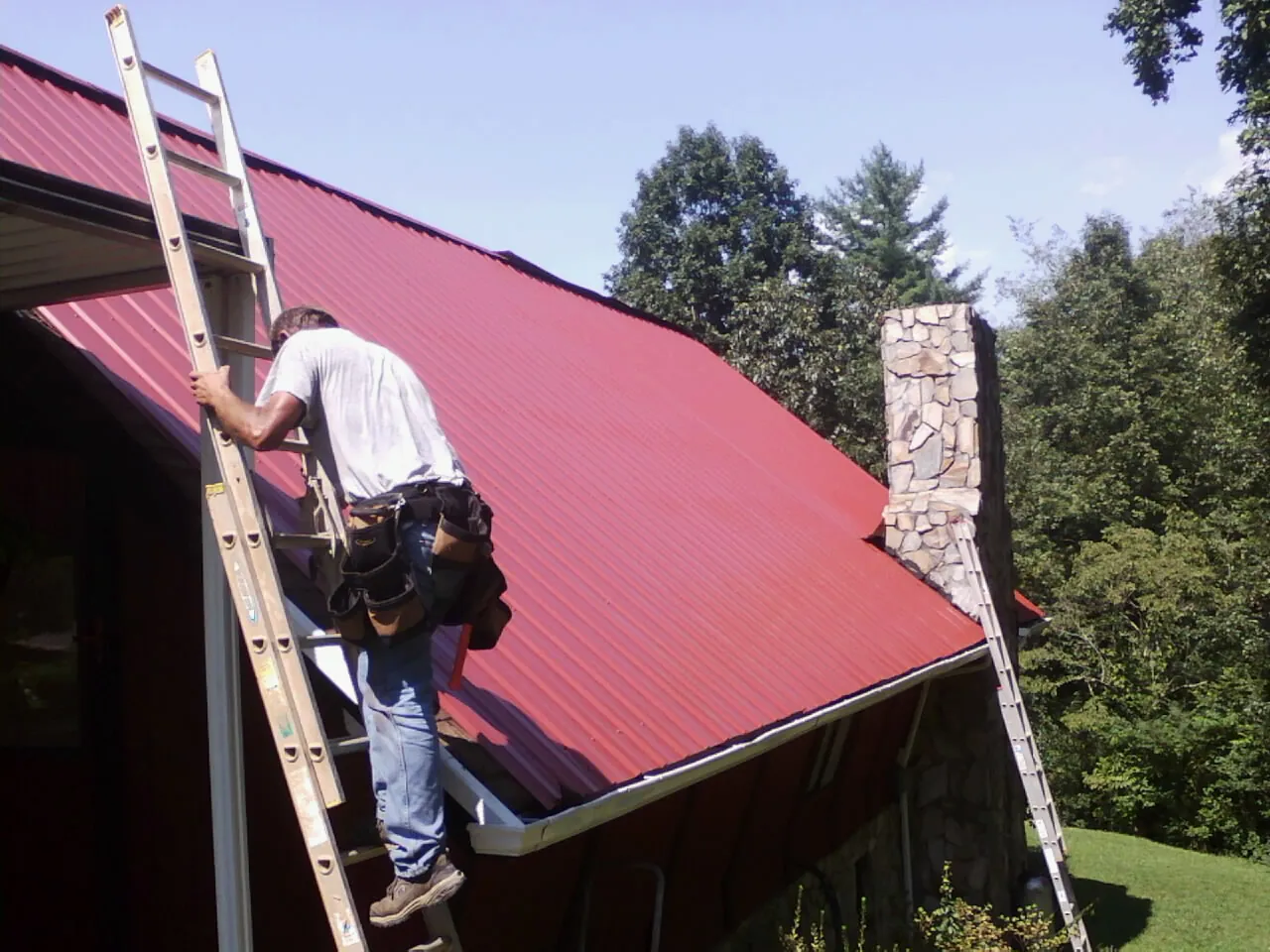 Roofer on a ladder beside a red metal roof and stone chimney — Soffit & Fascia Repair by Roofing Brothers in Creve Coeur